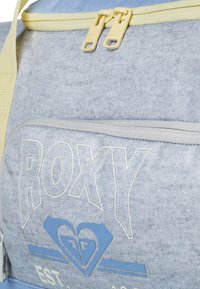 Sivý batoh so žltými zipsami a značkou Roxy. Má textúrovanú tkaninu, prednú kapsu s grafickým detailom srdca.