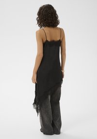 Robe nuisette noire avec bordure en dentelle, ourlet asymétrique, fines bretelles et texture satinée lisse. Portée par-dessus un jean ample gris.