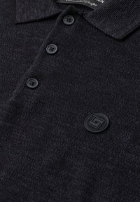 Polo en maille gris foncé avec trois boutons noirs et un emblème rond noir présentant un motif géométrique sur la poitrine.