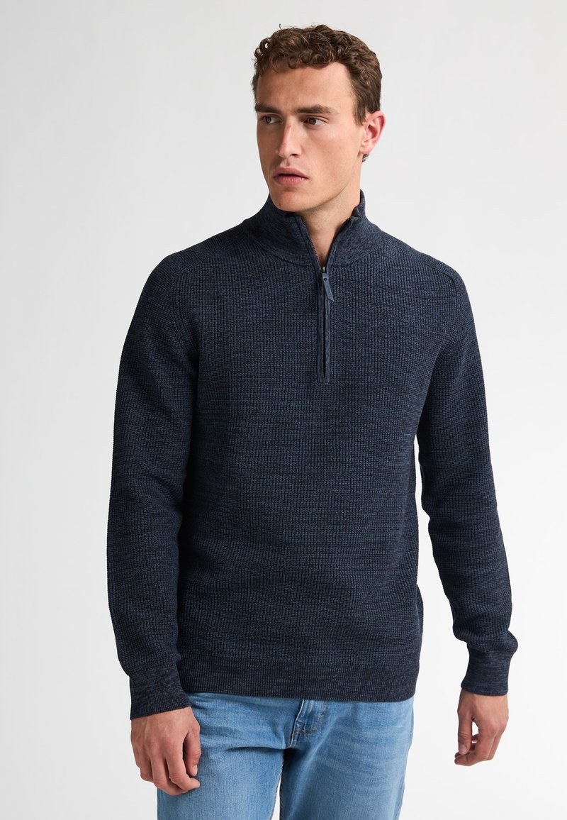 Navyblauwe zip-up trui met een gestructureerd breipatroon, lange mouwen en een hoge kraag, gecombineerd met lichtblauwe spijkerbroek.