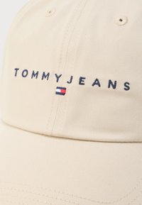 Μπεζ καπέλο με κεντημένο λογότυπο "TOMMYJEANS" σε σκούρο μπλε. Διαθέτει μικρή κόκκινη, λευκή και μπλε σημαία ως διακοσμητικό στοιχείο. Λείο υφασμάτινο υλικό.