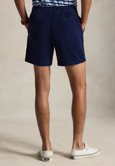Homme portant un short bleu marine avec une ceinture élastique et des baskets en toile blanches, debout les mains dans les poches, vue de dos.