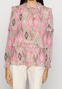 Femme portant un blouse rose à manches longues avec des motifs géométriques et des liens à franges, associée à un pantalon beige clair, montrée de la nuque aux hanches.