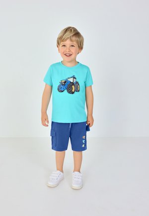 Ung dreng, der smiler, iført en turkis t-shirt med blå traktortryk, marineblå shorts med lapper og hvide sneakers, stående på hvidt gulv.