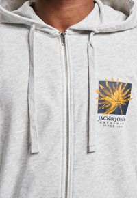 Grå hettejakke med glidelås og snor i hetta. Har et gult grafisk logo på brystet med "JACK & JONES" tekst. Myk tekstur.