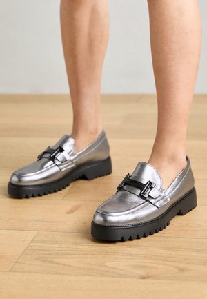 Metallische zilveren loafers met een zwarte accentgesp, dikke gestructureerde zool en een glad oppervlak. Beschikt over een ronde neus en een lage cut.