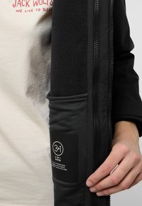 Giacca nera 3 in 1 con morbido rivestimento in pile, dotata di chiusura con zip, esterno testurizzato e etichetta informativa sulla tasca.