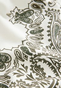 Valge kangas rohelise paisley-mustriga, milles on keerukaid kujundeid ja jooni, luues tekstuurse ja dekoratiivse pinna.