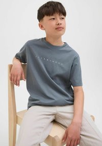 Camiseta gris de manga corta con el texto "JACK&JONES" en blanco en la parte frontal, acompañada de pantalones beige claros, sentado en una silla de madera.
