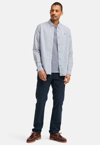 Geruit overhemd met lange mouwen in wit en marineblauw, gelaagd over een grijs T-shirt. Marineblauwe broek en bruine schoenen met veters en houten accenten.