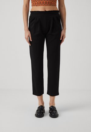 Kvinde iført sorte cropped bukser og sorte loafers med frynser, stående foran en ensfarvet lys grå baggrund.