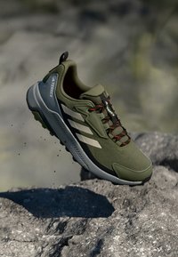 Olivgrüner Sportschuh aus Netzgewebe mit Gummisohle und roten Schnürsenkeln. Verfügt über drei weiße Streifen und eine Markenkennzeichnung an der Seite.