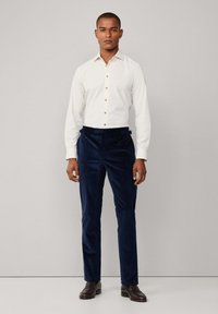 Witte overhemd met bruine knopen, gecombineerd met marineblauwe corduroybroek. Model staat rechtop en draagt donkerbruine schoenen.