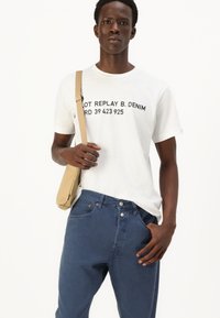 Witte t-shirt met zwarte tekst, gecombineerd met donkerblauwe jeans. Model houdt een tan schoudertas vast. Eenvoudig ontwerp, katoenen materiaal, losse pasvorm.