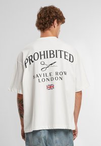 Valge üle mõõtme T-särk, millel on must trükk, mis ütleb "KEELATUD" koos käärid graafikaga, "SAVILE ROW LONDON" ja väike Briti lipp.