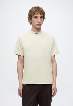Νέος άνδρας με σγουρά μαλλιά που φοράει γαλακτερό κοντομάνικο πουκάμισο με την ένδειξη «BOSS» στον γιακά και σκούρο παντελόνι σε απλό φόντο.