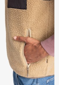 Beige Fleecejacke mit einer strukturierten Oberfläche, die eine seitliche Reißverschlusstasche und einen kontrastierenden schwarzen Patch aufweist. Der Ärmel zeigt einen rosa Bund.