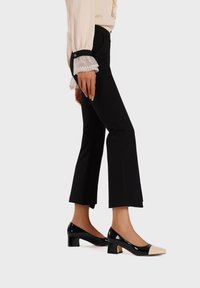 Tacchi in vernice nera con punta color crema, tacco basso a blocco e punta affusolata. Indossati con pantaloni neri aderenti e una blusa beige.