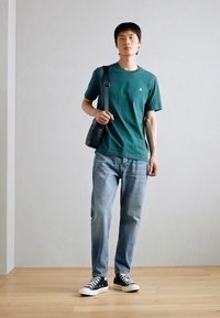 Green cotton t-shirt, light blue denim jeans, black and white canvas sneakers, and a small black shoulder bag. Light wooden floor.