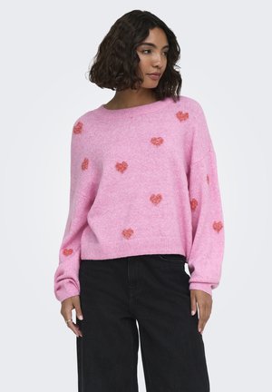Vrouw draagt een losse roze trui met rode hartpatronen en zwarte broek, staat met handen nabij haar heupen, kijkt iets opzij.