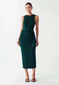 Robe longue verte en tissu lisse et extensible. Elle présente un corsage ajusté, une ceinture à la taille et une jupe droite.