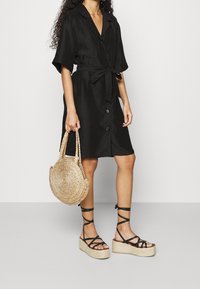 Donna che indossa un vestito nero con bottoni e cintura in vita, tiene una borsa di paglia intrecciata rotonda e indossa sandali platform neri con lacci.
