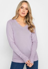 Violetti V-painikkainen neule, jossa on pitkät hihat, ribbireunat ja sileä, tiivis rakenne, yhdistettynä tummansinisiin farkkuihin.