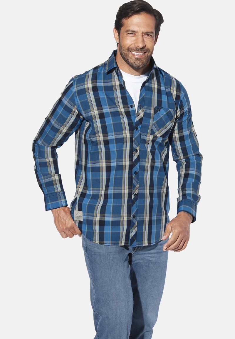 Lächelnder Mann mit Bart, der ein blaues karriertes Hemd über einem weißen T-Shirt und hellblauen Jeans trägt, vor einem weißen Hintergrund.