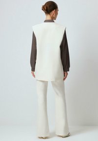 Chaleco sin mangas blanco sobre una camiseta de manga larga marrón oscuro, combinado con pantalones acampanados blancos y zapatos de tonos claros. Textura suave, ajuste a medida.