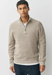 Beige Zippullover mit strukturiertem Strickdesign. Hat lange Ärmel und einen gerippten Saum, über einem weißen Oberteil getragen, kombiniert mit dunklen Jeans.