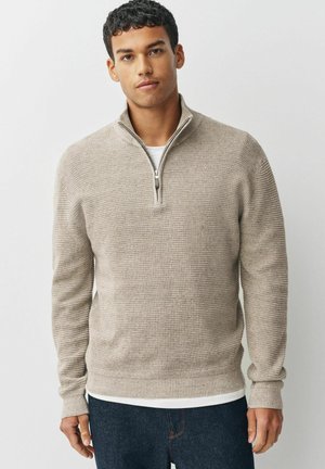 Beige zippered sweater with a textured knit design. Features long sleeves and a ribbed hem, layered over a white top, paired with dark jeans.