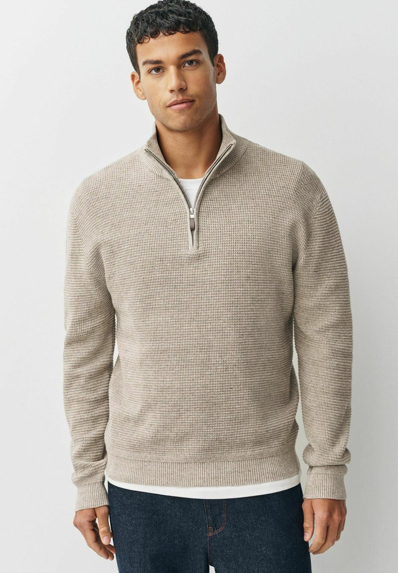 Beige Zippullover mit strukturiertem Strickdesign. Hat lange Ärmel und einen gerippten Saum, über einem weißen Oberteil getragen, kombiniert mit dunklen Jeans.