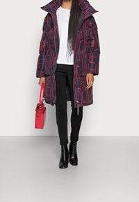Abrigo largo acolchado en azul marino y burdeos con un diseño de letras atrevidas, que presenta un collar alto y cierre de cremallera, combinado con pantalones negros y un bolso rojo.