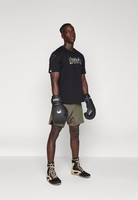 Black athletic T-shirt with colorful logo, olive shorts, black boxing gloves, and black/gold training shoes. Model stands facing forward.