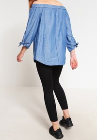 Kvinna i axelbandslös blå blus med knutna ärmar, svarta leggings och svarta sneakers, stående mot en enkel ljus bakgrund.