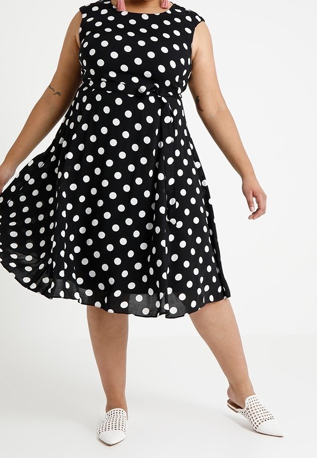 Vestido negro con lunares, con la cintura atada, diseño sin mangas y falda acampanada. Combinado con zapatos blancos sin cordones. Tejido texturizado.