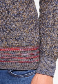 Gestrickter Pullover in Blau und Braun mit strukturiertem Muster, ausgestattet mit einem rot gestreiften Saum und einem kleinen Markenschild am unteren Rand.