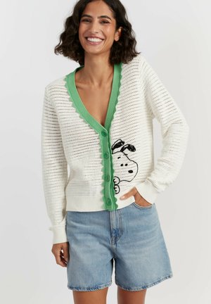 Donna sorridente che indossa un cardigan bianco lavorato a maglia con rifiniture verdi e motivo Snoopy, abbinato a pantaloncini di jeans azzurri, posa con la mano in tasca.