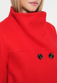 Cappotto rosso di lana con colletto alto, dotato di due bottoni neri e una superficie liscia. Il design mette in risalto il calore e il colore vivace.