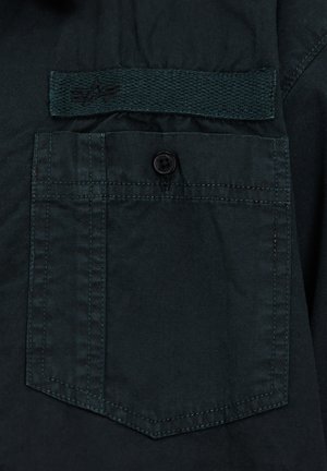 Tela de camisa en verde oscuro con un bolsillo trasero con cierre de botón y una etiqueta tejida sobre el bolsillo. Textura suave con costuras de contraste.