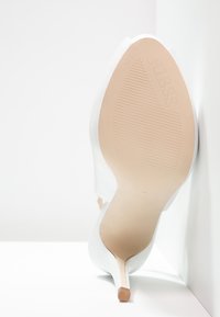 Scarpa bianca con tacco alto e punta affusolata, dalla superficie liscia, e suola in gomma beige con il marchio "GUESS" impressa sul fondo.