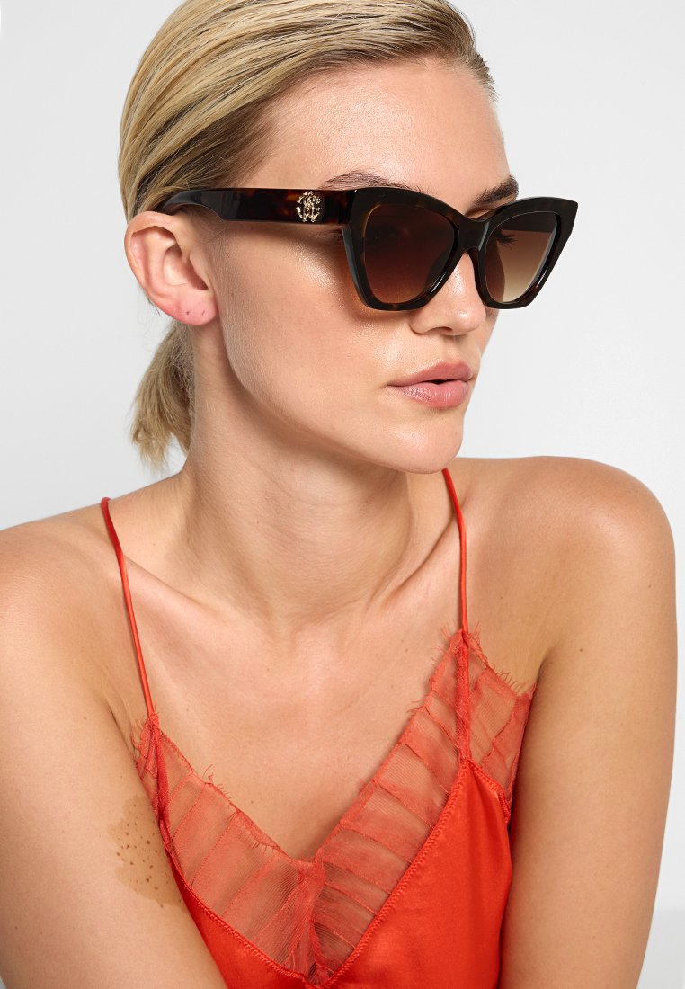 Des lunettes de soleil en œil de chat avec une monture en écaille sombre, des verres dégradés marron et un accent en or avec le logo sur le côté. Fini lisse et brillant.