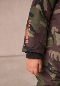 Kamuflāžas drēbes ziemas apstākļiem, zaļās un brūnās krāsās, ar SUPRDRY logo oranžā krāsā, ar ribotu melnu apkakli.