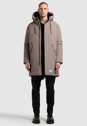 Parka beige acolchado con capucha, que cuenta con cierre de cremallera y dos bolsillos laterales. Unos pantalones negros y botas con cordones complementan el conjunto.