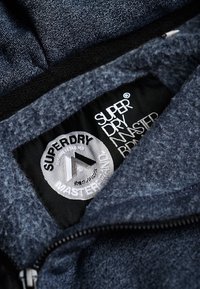 Granatowy materiał polaru z czarną metką ozdobioną napisem "SUPERDRY" i logo. Zbliżenie na wewnętrzną podszewkę i detal zamka.