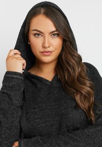 Mustapaita, jossa on huppu ja teksturoitu kangas, V-mallinen kaula-aukko, nyörit ja kontrastiväriset mustat korostusnauhat sivuilla.