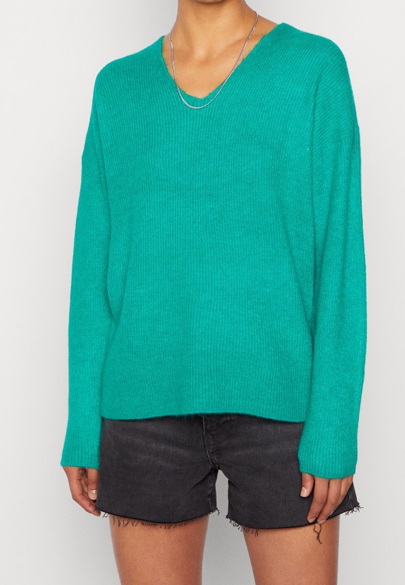 Personne portant un pull col V turquoise vif et un short en jean noir effiloché, avec un collier chaîne en argent.