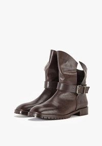 Par de botas de cuero marrón hasta el tobillo con correas de hebilla lateral y tacones bajos en un fondo blanco.