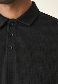 Sort polo t-shirt med tekstureret stof, tre knapstil og en struktureret krave. Materialet har et diskret mønster.