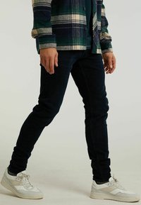 Jeans oscuros de corte ajustado combinados con una camisa de franela a cuadros en verde, marino y crema, completados con zapatillas blancas.